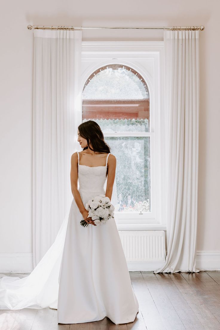 a woman standing in front of a window wearing a white dress and holding a bouquet