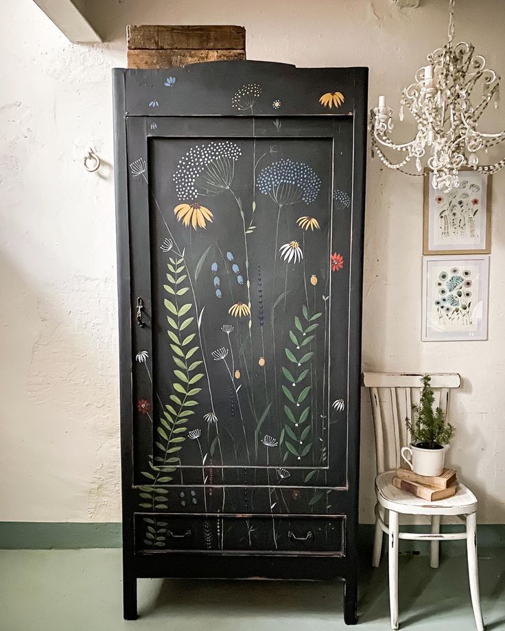 a black cabinet with flowers painted on the front and sides, next to a white chair