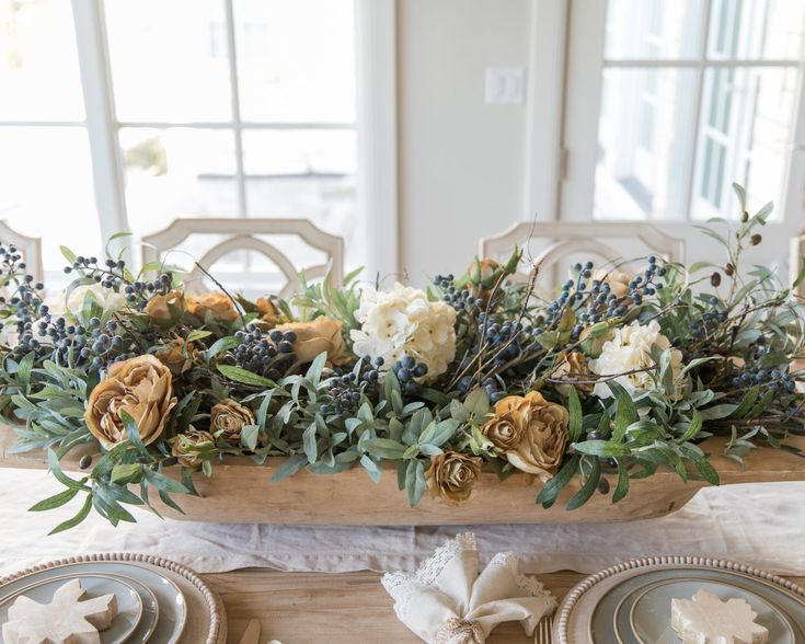 an arrangement of flowers and greenery sits on a dining room table with place settings