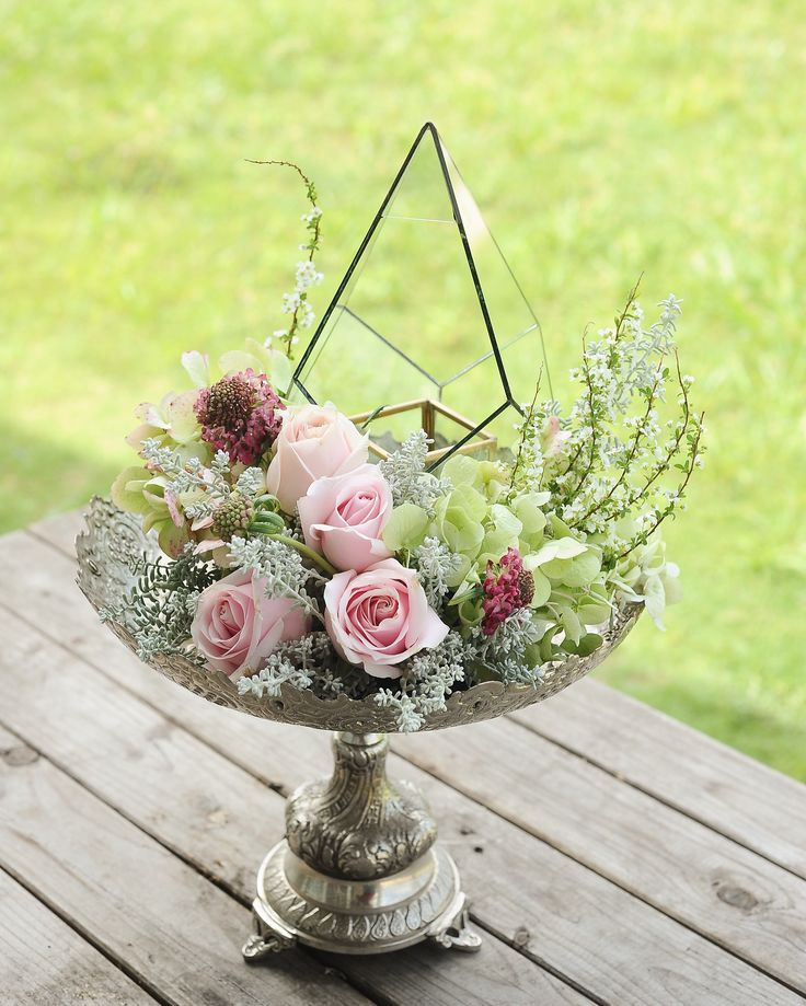 a flower arrangement in a metal bowl on a wooden table with grass and trees behind it