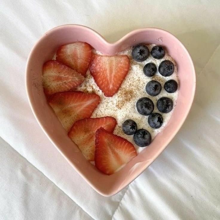 a heart shaped bowl filled with fruit on top of a white bed sheet in the shape of a heart