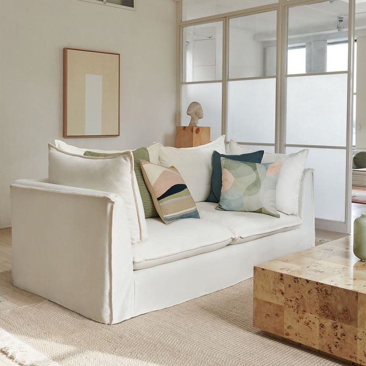 a white couch sitting in front of a window next to a table with a vase on it