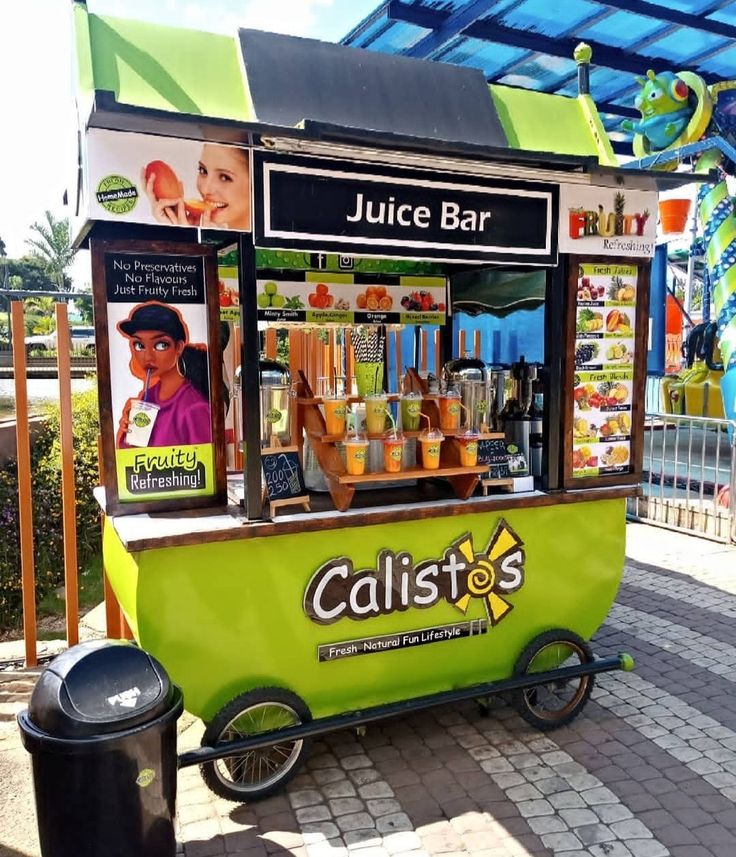 a green juice cart sitting on top of a brick sidewalk next to a trash can