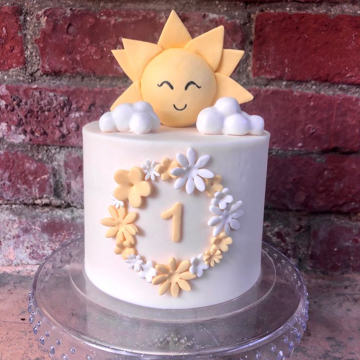 a white cake decorated with flowers and a sun on top is sitting on a clear platter