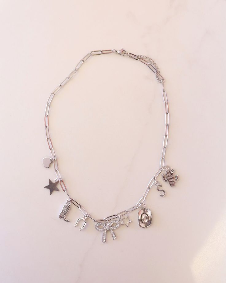 Introducing our Glamour Cowgirl Charm Necklace: a stylish statement piece crafted from durable stainless steel. Adorned with Western-inspired charms, this 17-inch necklace exudes cowboy chic with every wear. Perfect for adding a touch of glamour to your cowgirl ensemble. Let us know at checkout what letter you'd like us to replace the "S" charm with! If you don't leave a note, it will be shipped with the "S" charm. Since stainless steel is a lower grade metal, it can tarnish if exposed to harsh Silver Charm Necklace Aesthetic, Western Charm Necklace, Silver Necklaces With Charms, Glamour Cowgirl, Cowgirl Necklace, College Crafts, Cowboy Jewelry, Cowgirl Necklaces, Cowboy Chic