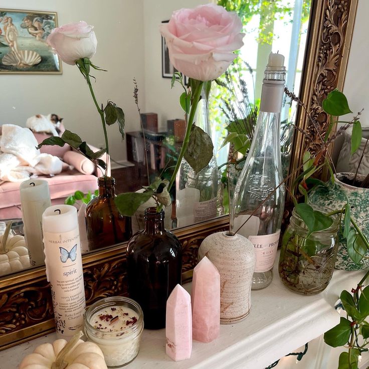 a mantle with candles, bottles and flowers on it in front of a mirror that is reflecting the room