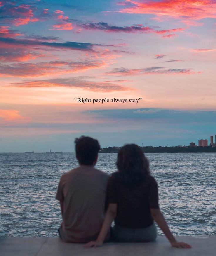 two people sitting on the edge of a pier looking out at the ocean with a sunset in the background