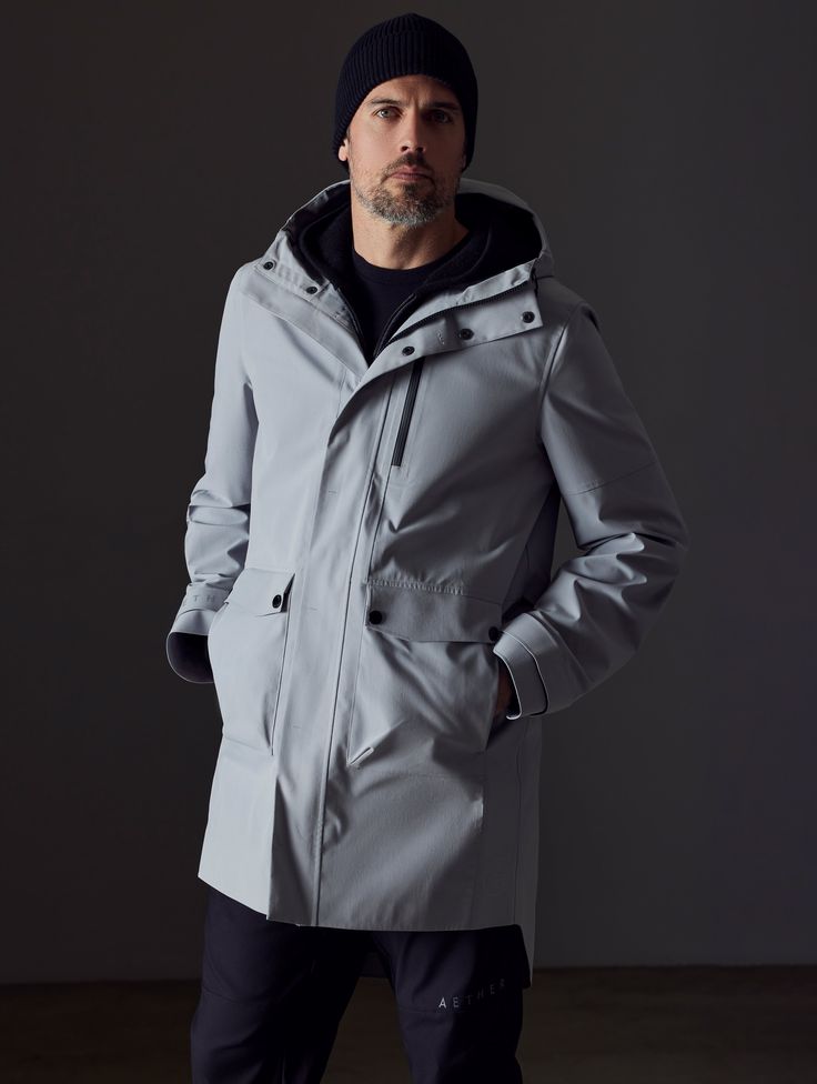 Front body view of man wearing Anders Rain Jacket in Sky Grey from AETHER Apparel. Modern Waterproof Raincoat For Workwear, Modern Raincoat With Detachable Hood And Long Sleeves, Weatherproof Long Coat For Rainy Weather, Weatherproof Long Coat For Workwear, Waterproof Long Coat For Work, Waterproof Long Coat For Workwear, Techwear Outerwear With Detachable Hood For Work, Modern Nylon Outerwear For Rainy Weather, Modern Waterproof Outerwear For Outdoor