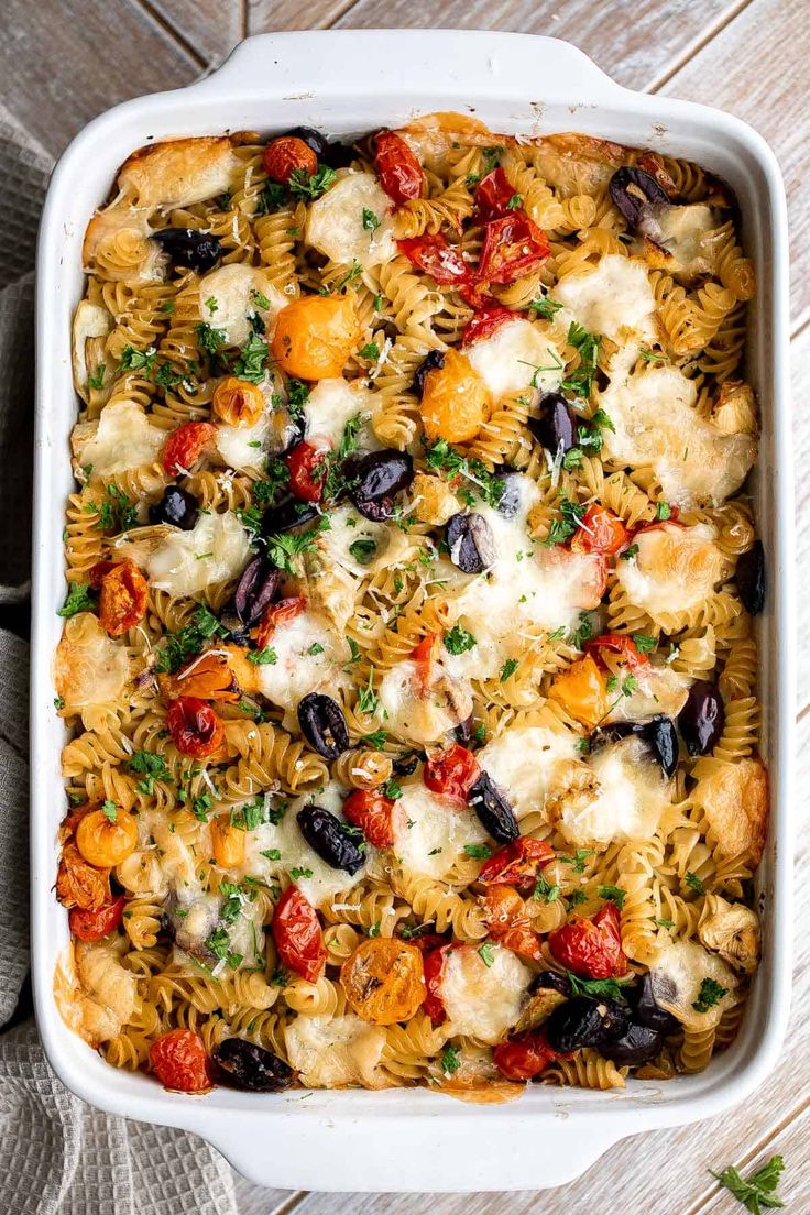 a white casserole dish filled with pasta, olives, tomatoes and mozzarella