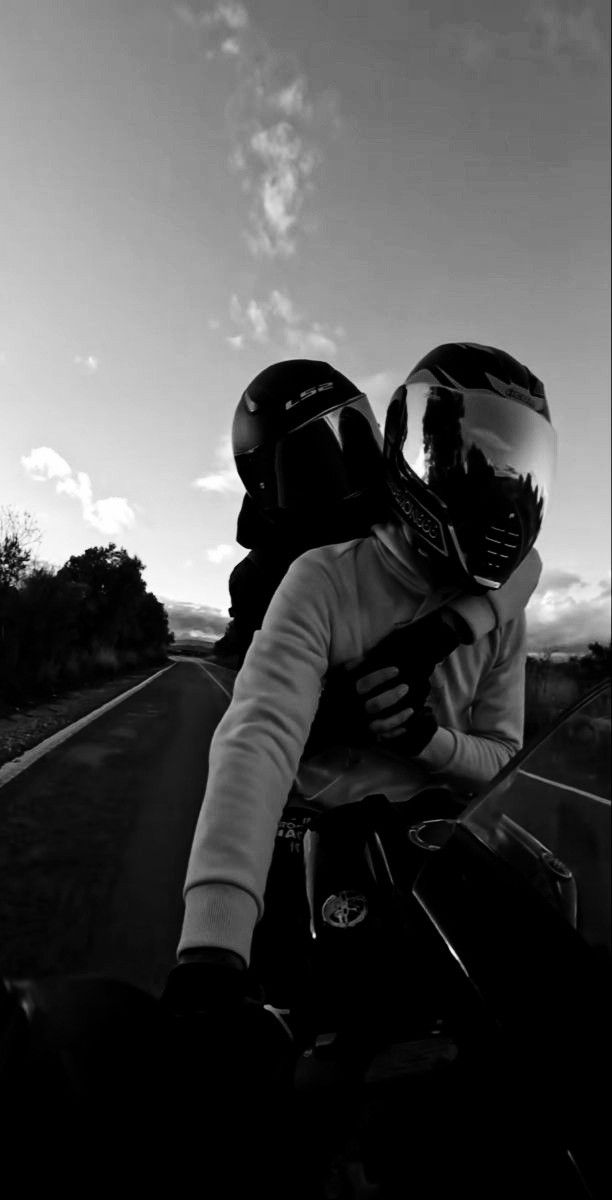 two people riding on the back of a motorcycle down a country road in black and white