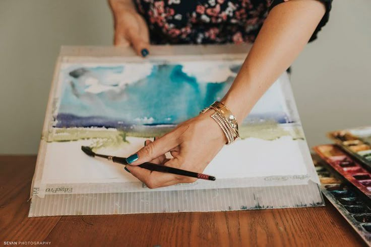 a woman is holding a paintbrush and painting on a piece of paper with watercolors