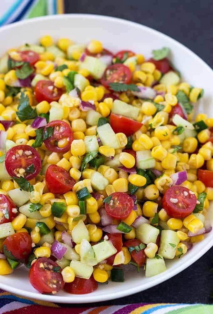 a white bowl filled with corn, tomatoes and cucumber on top of a colorful table cloth