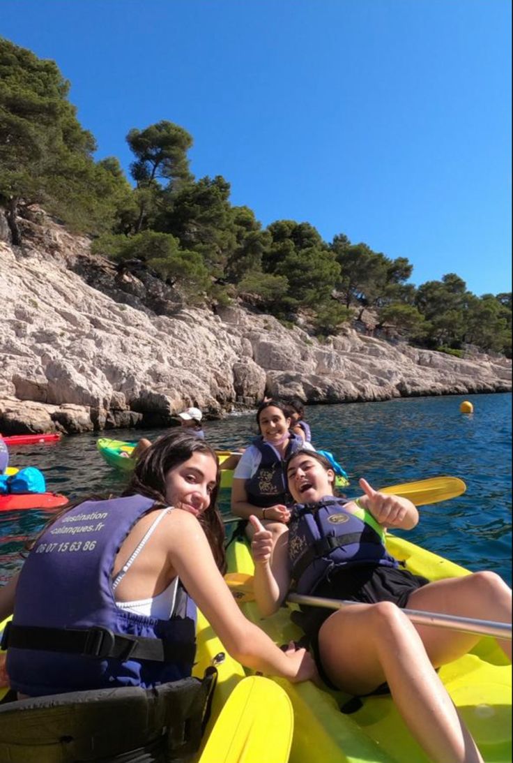 three people in kayaks paddling on the water near rocks and trees, with one person smiling at the camera