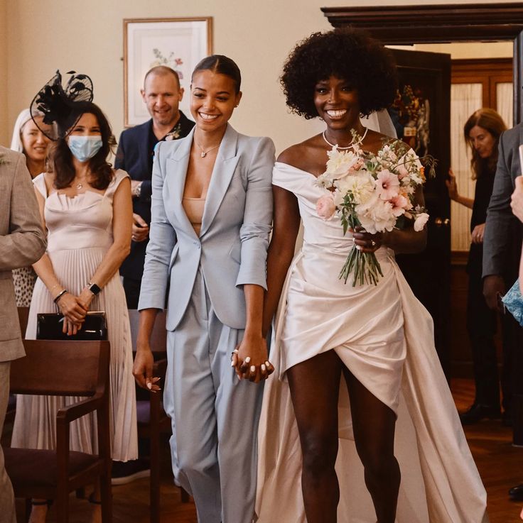 two women in wedding attire walking down the aisle with people behind them wearing face masks