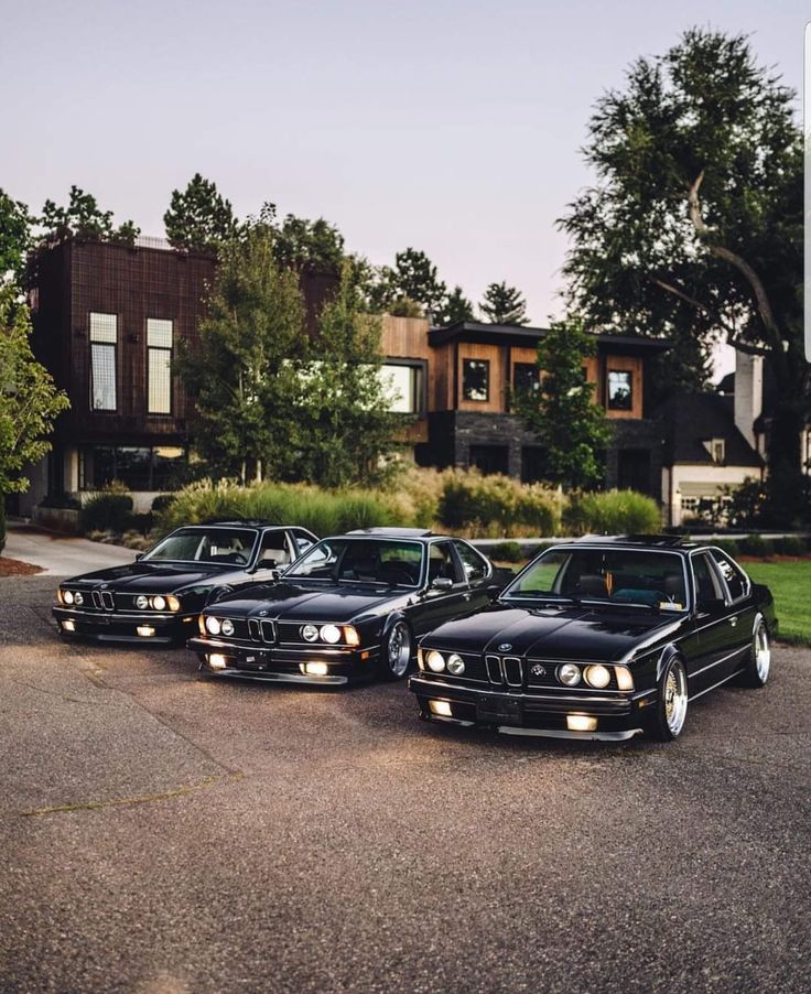 three black bmws are parked in front of a house with trees on the other side