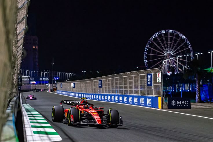 a man driving a red car down a race track at night with a ferris wheel in the background