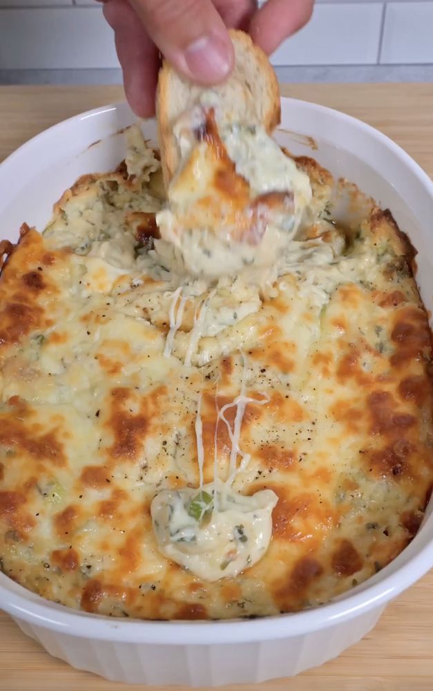 a person dipping cheese into a casserole in a white dish on top of a wooden table