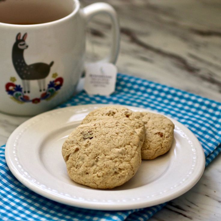 two cookies on a white plate next to a cup of tea and a blue checkered napkin