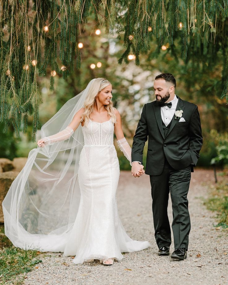 a bride and groom holding hands while walking down a path with lights strung from the trees