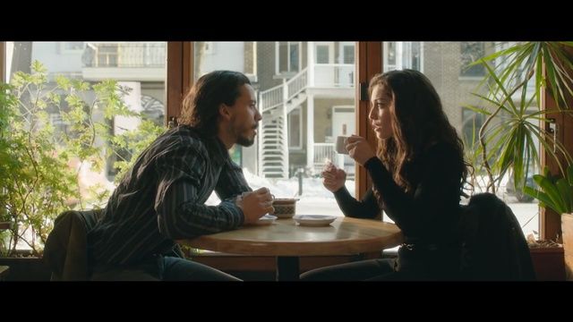 a man and woman sitting at a table in front of a window talking to each other