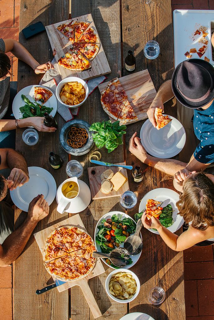 people sitting at a table eating pizza and drinking wine, with one person holding a slice of pizza