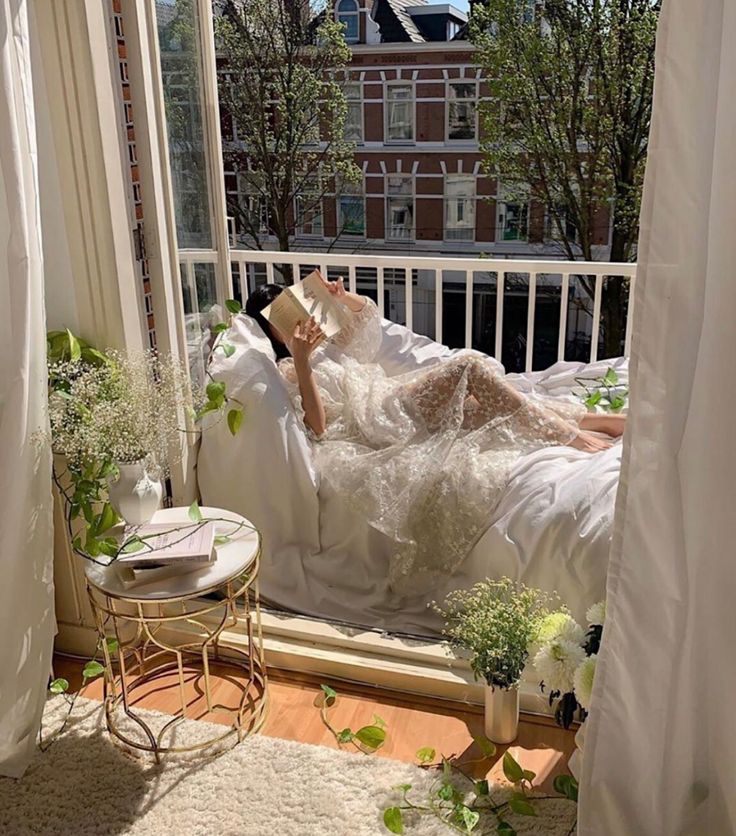 a woman laying in bed on top of a white blanket next to a window with curtains