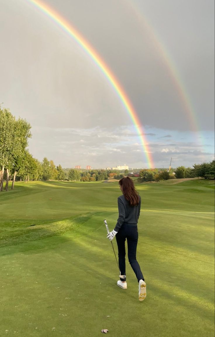 a woman is playing golf with a rainbow in the sky above her and behind her