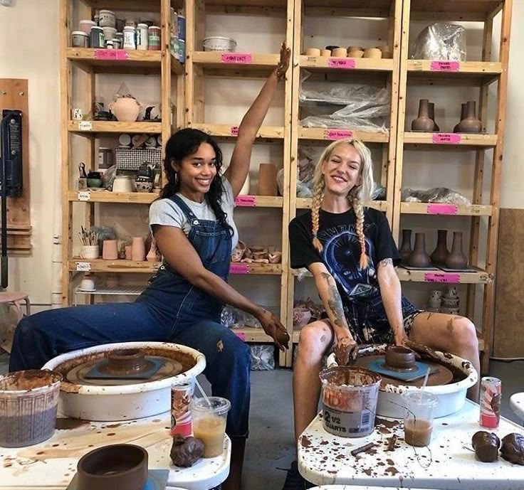 two women are sitting on buckets in a pottery shop and one is holding up her hand