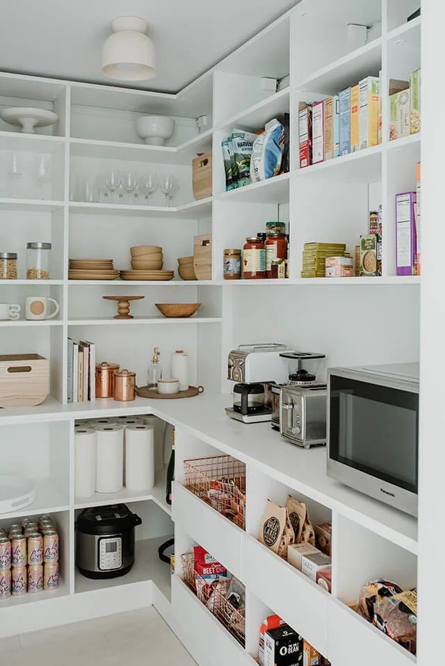 a kitchen with white shelving and lots of food on the counter top in front of a microwave