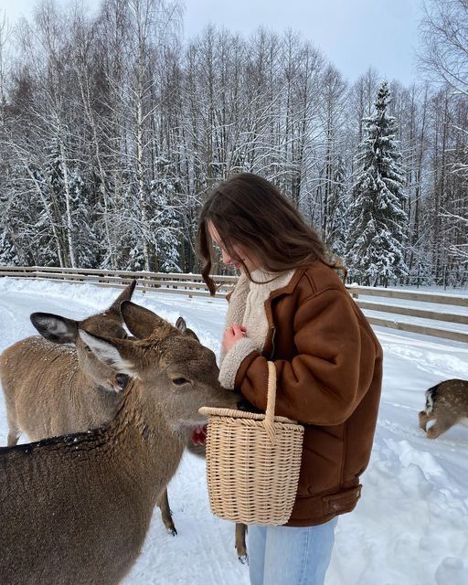 a woman feeding two deer in the snow with a basket on her shoulder and another deer standing next to her