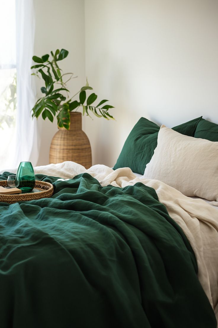 a bed with green sheets and pillows in front of a potted plant on the floor