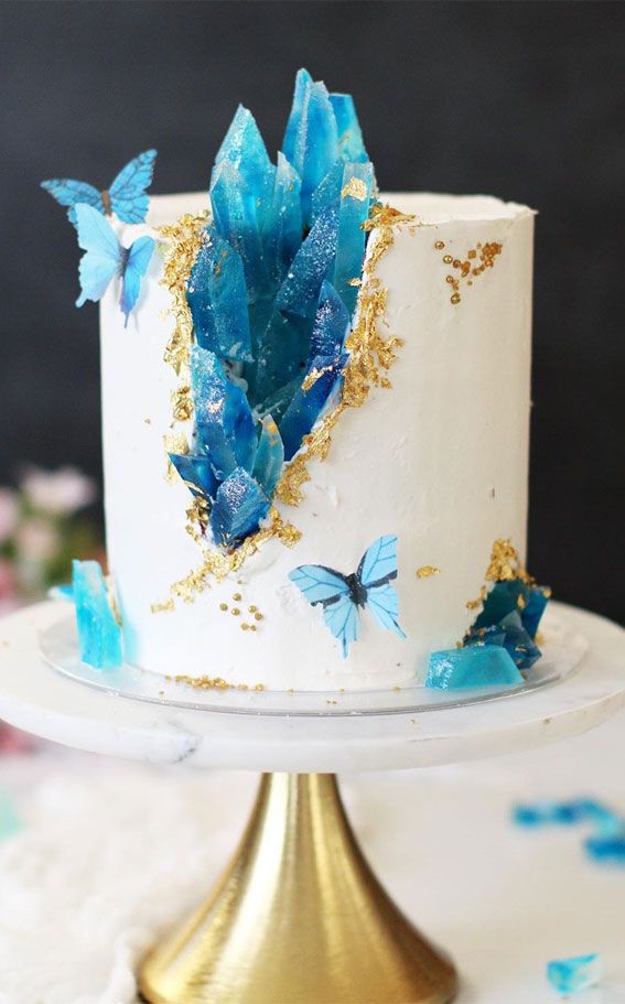 a white cake with blue butterflies on top and gold trim around the edges is sitting on a stand