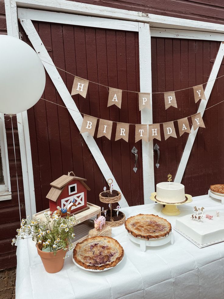 This photo contains a variety of pies and cupcakes as well as 2 white cakes across a white table as well as mini animal figures such as a pig, horse, cow, rooster, and a farm house to tie it all together. There are also balloons the color white, red and cow print on both ends of the table. Farm Birthday Party Aesthetic, Simple Farm Birthday, Modern Farm Theme Birthday, Vintage Farm Birthday Party, Neutral Farm Birthday Party, Minimalist Farm Birthday Party, Neutral Barnyard Birthday, Farm Theme Centerpieces, Barnyard Dance Birthday Party