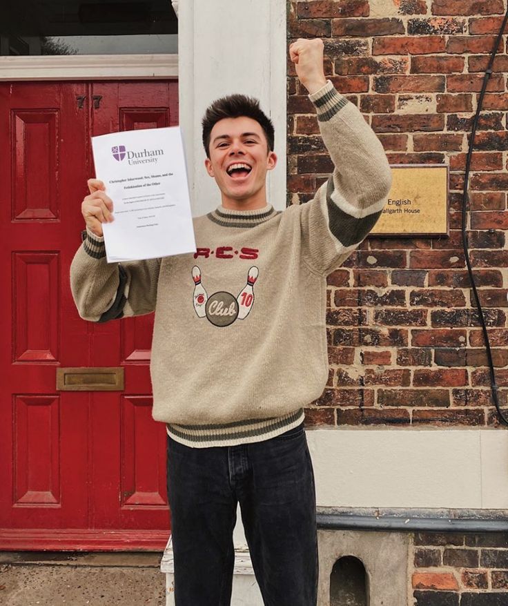 a young man holding up a paper in front of a red door with his fist raised