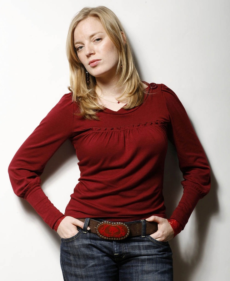 a woman standing with her hands on her hips wearing a red shirt and jeans, looking at the camera