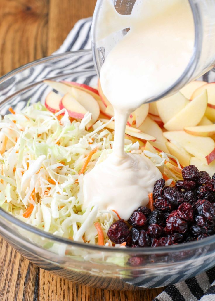 someone pouring dressing into a bowl filled with apples and cranberry slaw salad