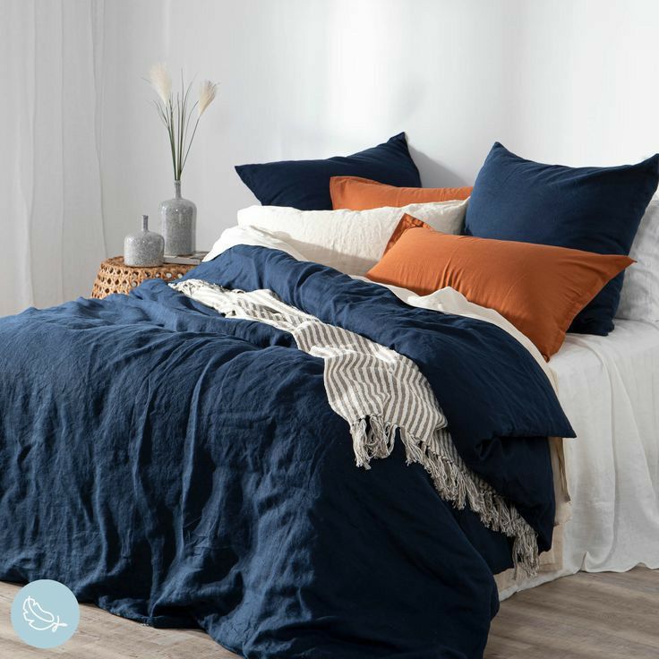 a bed with blue linens and orange pillows in a white walled room next to a wicker basket