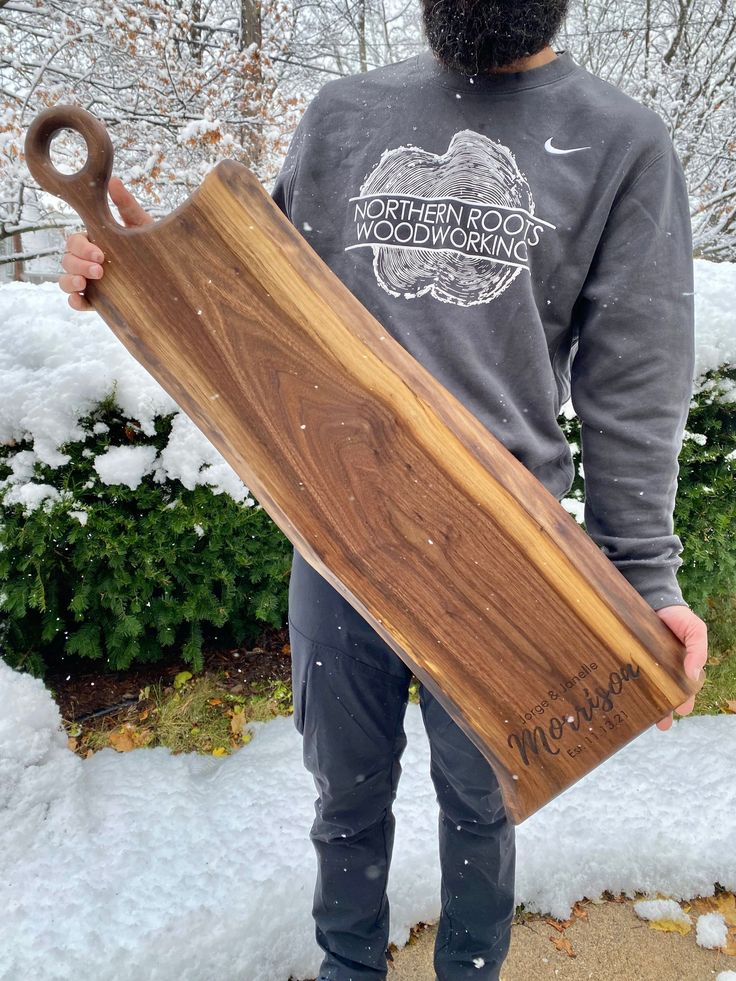 a man holding a long wooden skateboard in his hands and wearing a beard with snow on the ground behind him