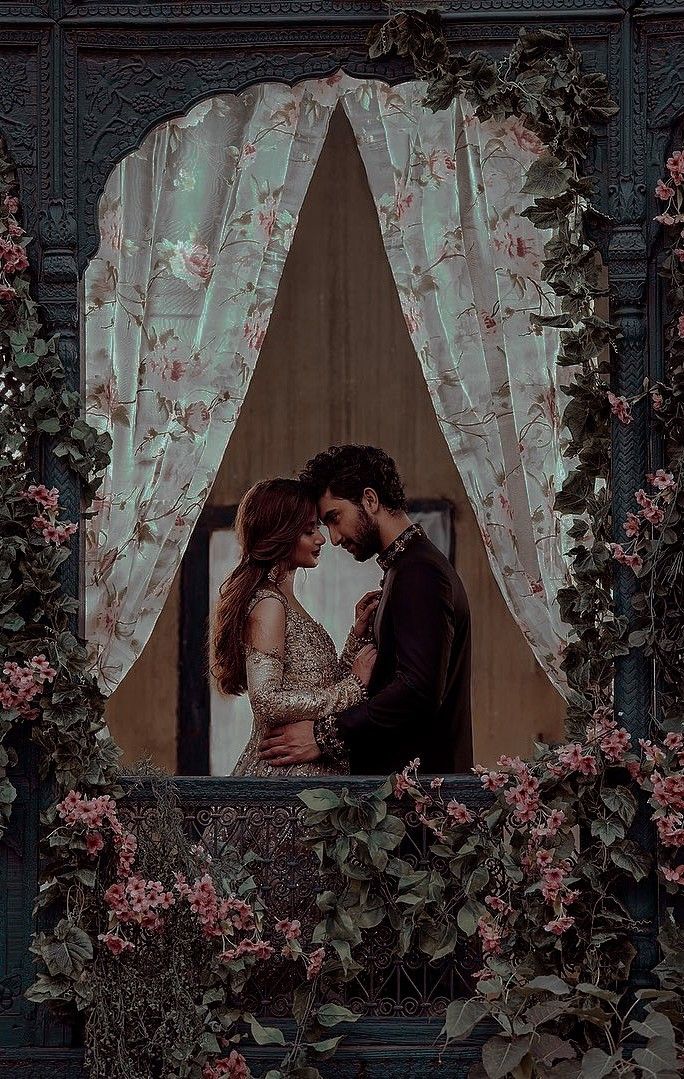 a man and woman standing next to each other in front of a window covered with flowers