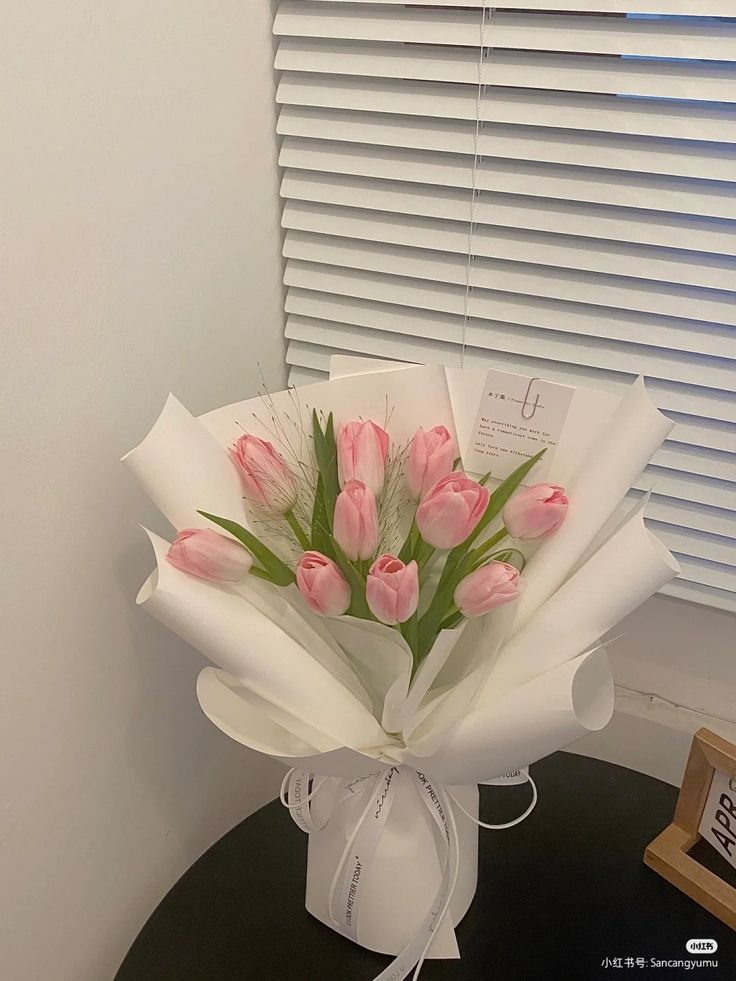 a bouquet of pink tulips wrapped in white paper on a table next to a window