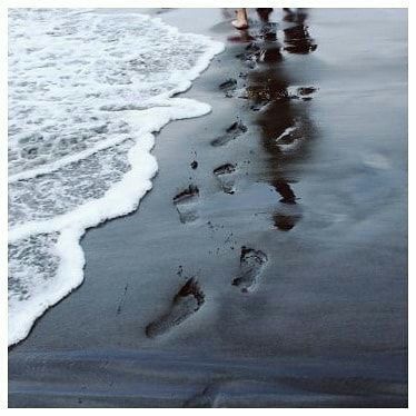 two people walking on the beach with footprints in the sand and waves coming toward them