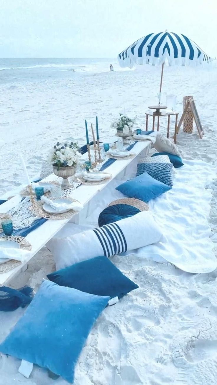 a long table set up on the beach with blue and white pillows, place settings and an umbrella