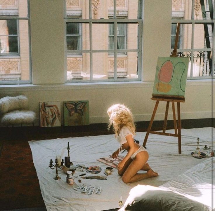 a woman sitting on top of a bed next to an easel