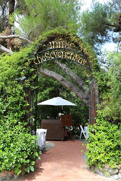 the entrance to an outdoor restaurant surrounded by greenery and trees, with umbrellas over tables