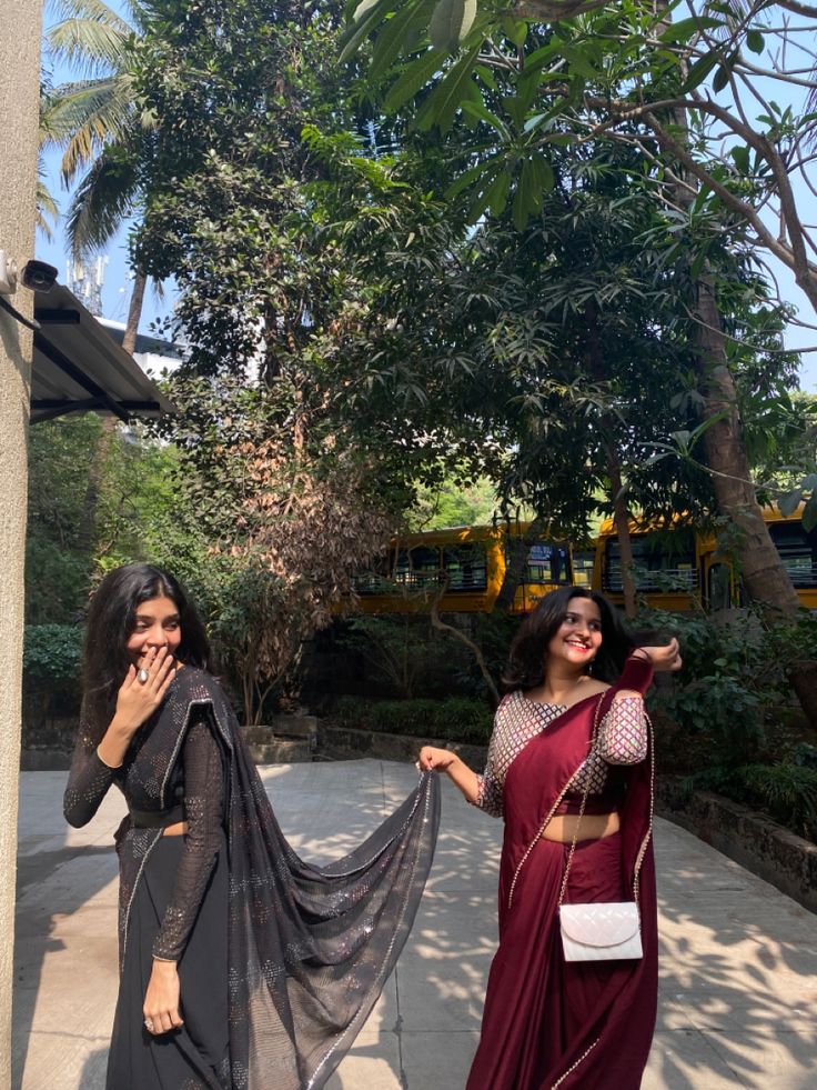 two women standing next to each other in front of some trees and bushes, one wearing a sari