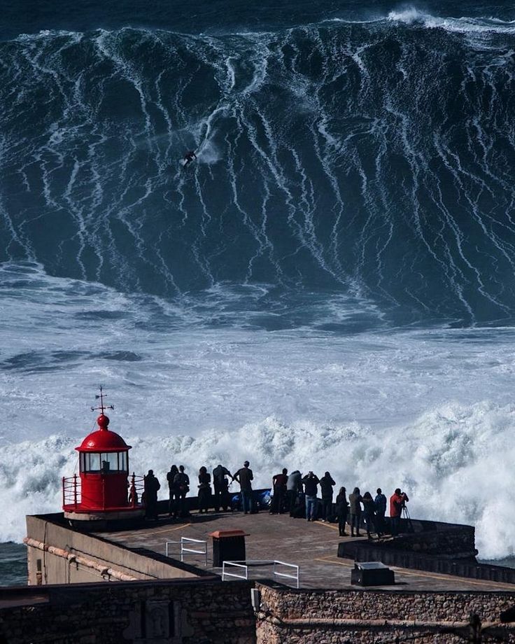 a group of people standing on top of a pier next to a large wave in the ocean