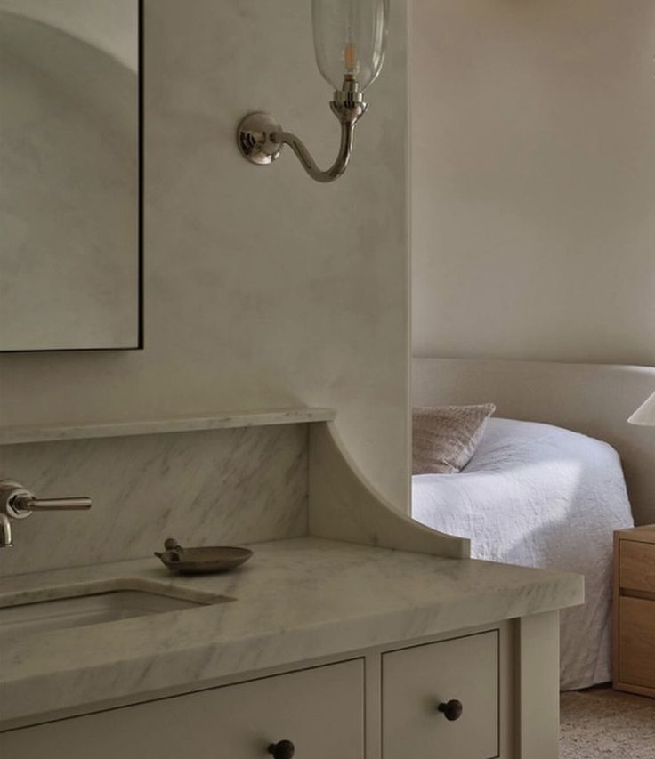 a bathroom sink sitting under a mirror next to a white bed and dresser with drawers