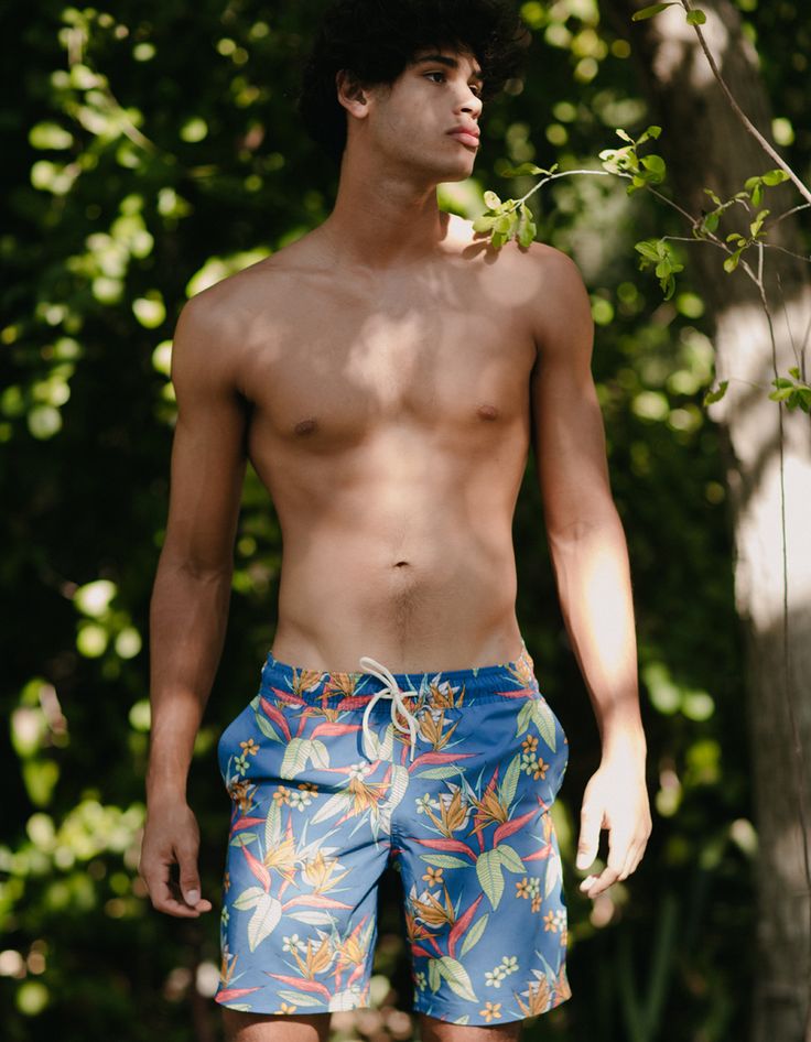 a shirtless young man standing in front of some trees and bushes wearing blue swim trunks