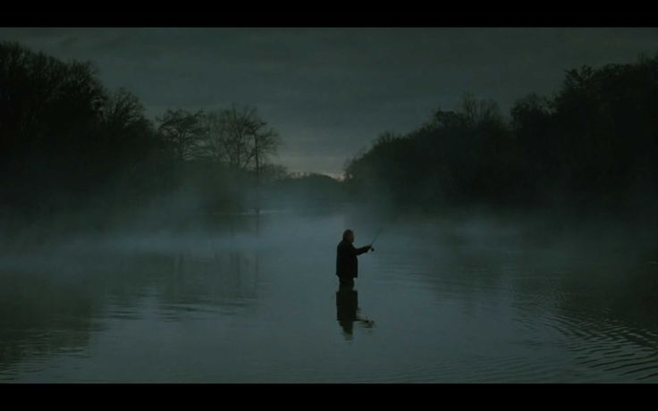 a man standing in the middle of a body of water surrounded by fog and trees