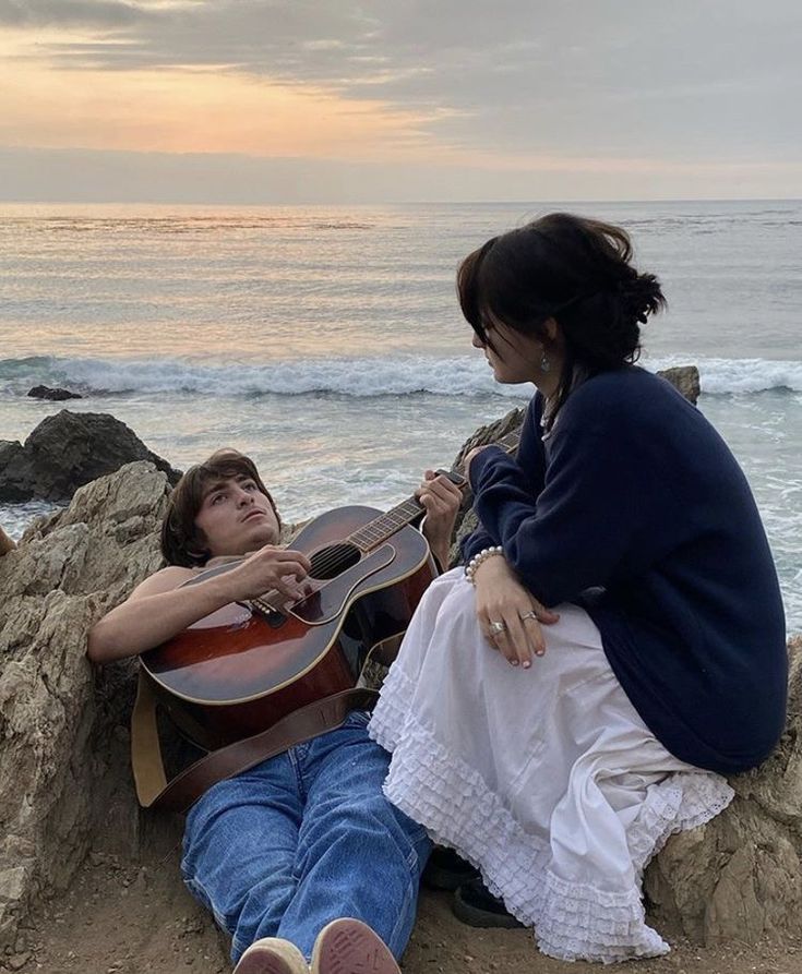 a woman sitting next to a man on top of a beach holding an acoustic guitar